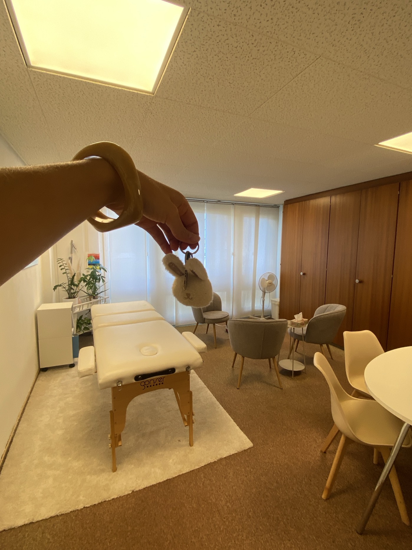 Alexane tend la clé de son cabinet de massage à Morges : table de massage blanche sur un tapis moelleux, fauteuils et plantes vertes